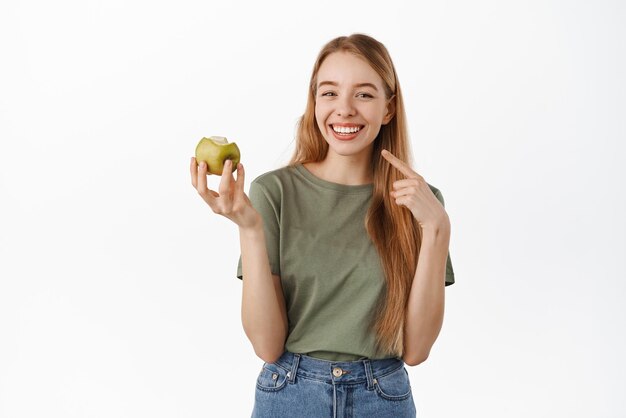 白い背景に立っている白く健康な歯を示す彼女の白い完璧な笑顔を指して青リンゴを食べる幸せな若い女性