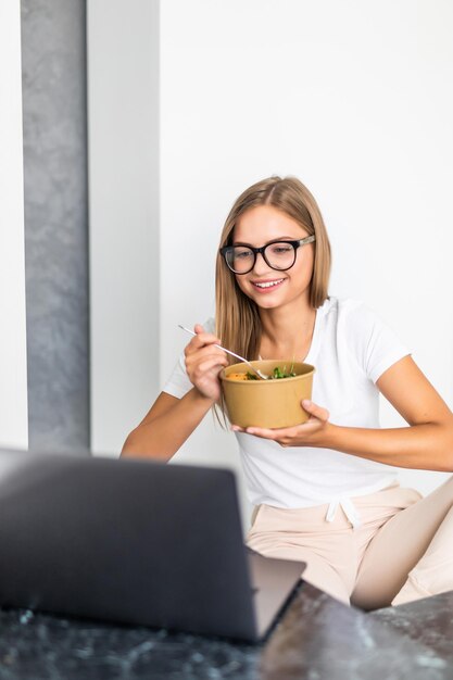 Happy young woman eating salad from a bowl while standing on a kitchen and watching movie on laptop
