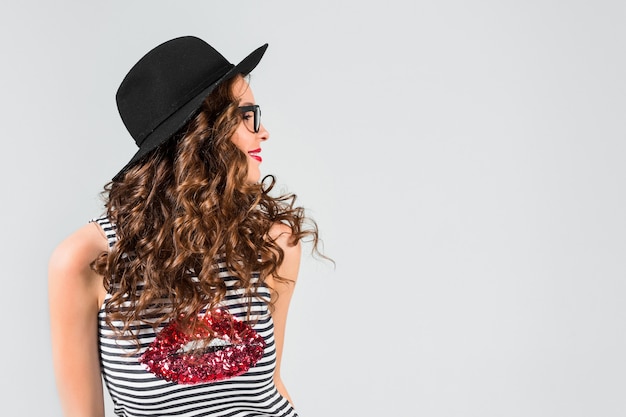 Happy young woman in glasses and hat on gray studio wall