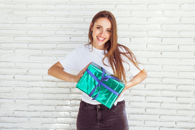 Happy young woman holding gift box in front of brick wall