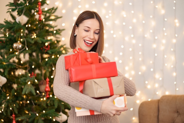 Happy young woman holding many present boxes with Christmas tree and lights