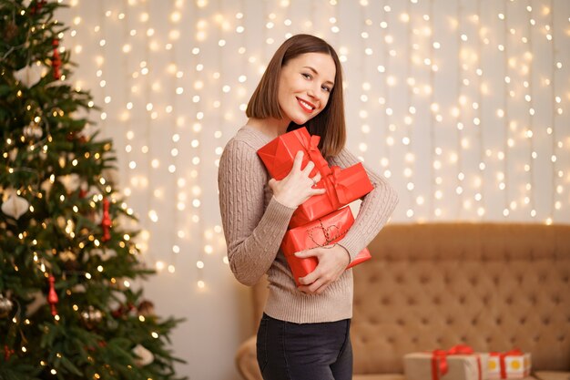 Happy young woman holding many present boxes with Christmas tree and lights