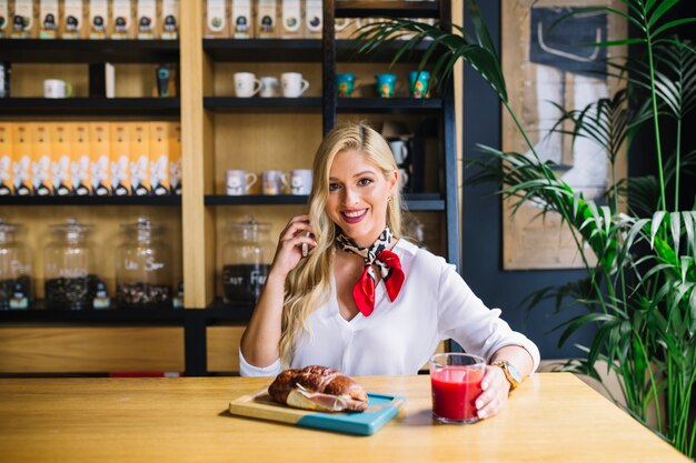 Happy young woman holding mobile phone in hand sitting at desk with baked bread and juice