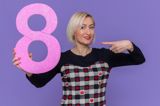 happy young woman holding number eight pointing with index finger at it