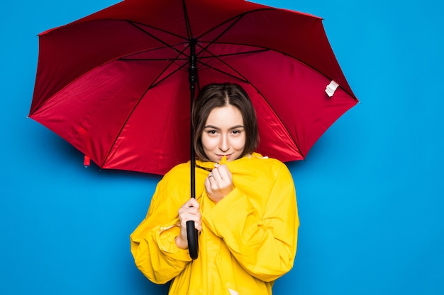 Happy young woman holding umbrella with yellow raincoat and blue wall