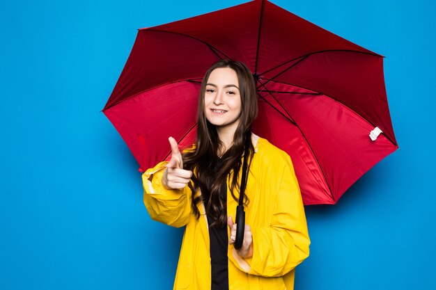 Happy young woman holding umbrella with yellow raincoat and blue wall