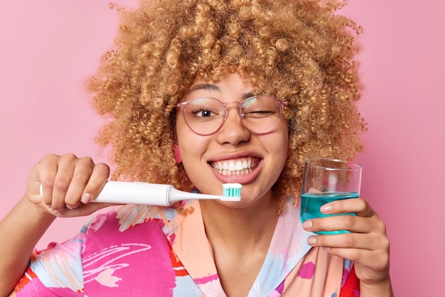 Бесплатное фото Счастливая молодая женщина держит стакан свежей жидкости для полоскания рта и электрическую зубную щетку, подмигивает глазу, подвергается гигиеническим процедурам, носит очки и повседневную рубашку, изолированную на розовом фоне. концепция ухода за полостью рта