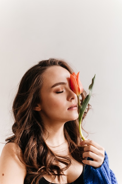 Happy young woman holds tulip. Brunette curly girl in black tee poses on white isolated background.