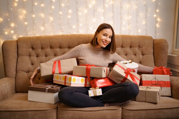 Happy young woman hugging many present boxes sitting crossed legs on a camel sofa with with lights