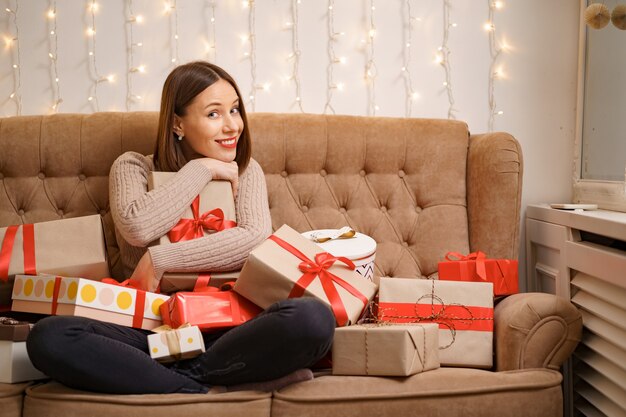 Happy young woman hugging many present boxes sitting crossed legs on a camel sofa with with lights