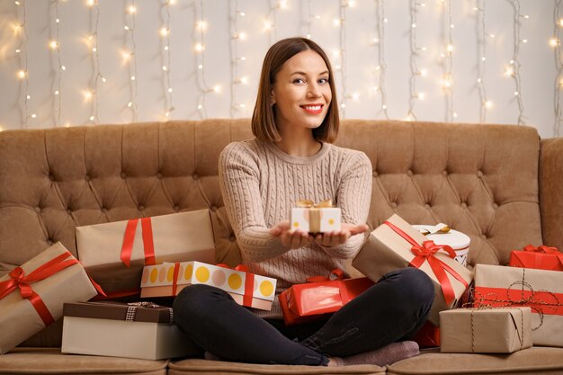 Happy young woman hugging many present boxes sitting crossed legs on a camel sofa with with lights