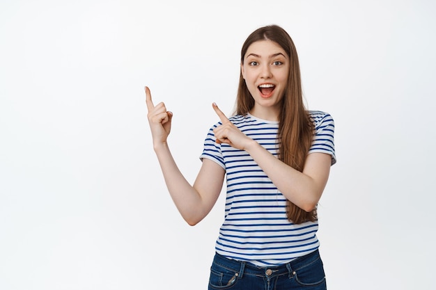 Happy young woman inviting and showing product, pointing fingers at upper left corner, smiling amazed on white