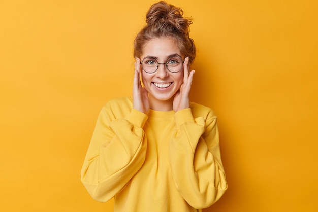Happy young woman keeps hands on face smiles broadly shows white teeth wears round glasses for sight casual jumper isolated over yellow background. Human face expressions and emotions concept