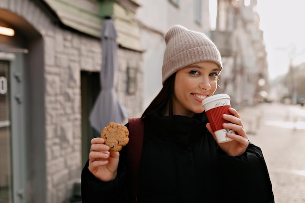Счастливая молодая женщина в вязаной шапке и темной куртке на улице с кофе на вынос и печеньем Обаятельная активная девушка отдыхает в городе в солнечный день
