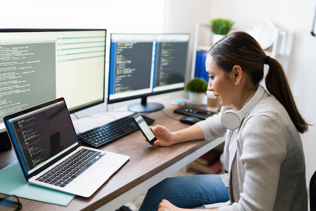 Happy young woman looking at her smartphone and texting while coding on her computer. Programmer working on developing software at home