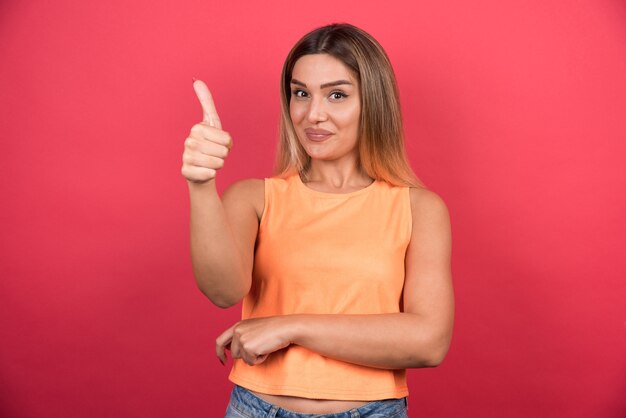 Happy young woman making thumbs up on red wall. 