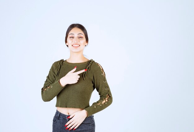 A happy young woman model standing and pointing at herself over white wall.