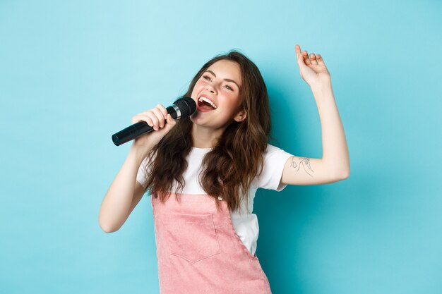 Happy young woman perform song, singer holding microphone, dancing and singing at karaoke, standing over blue background