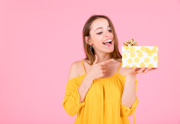 Happy young woman pointing finger to gift box against pink background