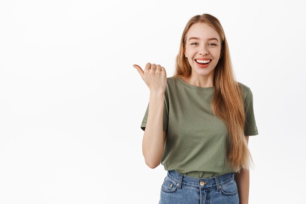 左を指して笑って、完璧な白い笑顔を見せて、白い壁にTシャツとジーンズで立っている幸せな若い女性