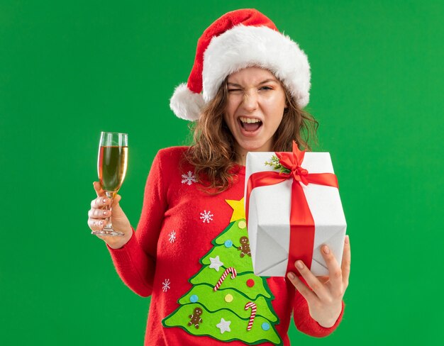 Happy young woman in red christmas sweater  and santa hat holding glass of champagne and present  looking at camera winking and  smiling cheerfully standing over green  background