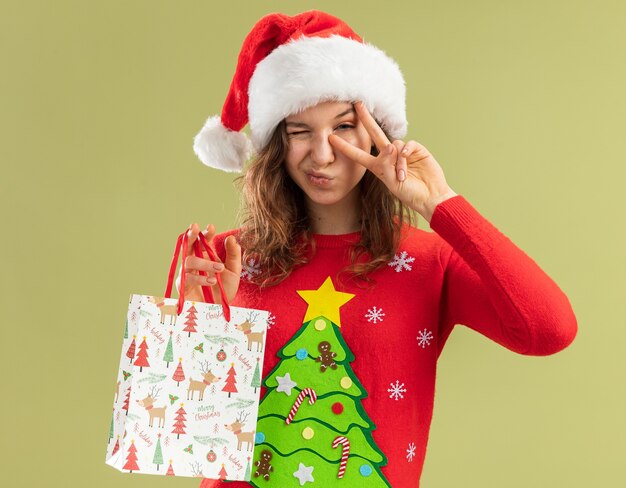 빨간 크리스마스 스웨터와 산타 모자 녹색 벽 위에 v 기호 서 보여주는 윙크 크리스마스 선물 종이 가방을 들고 행복 한 젊은 여자