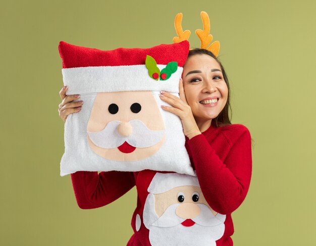 Счастливая молодая женщина в красном рождественском свитере в забавной оправе с оленьими рогами держит рождественскую подушку, весело улыбаясь, стоя над зеленой стеной