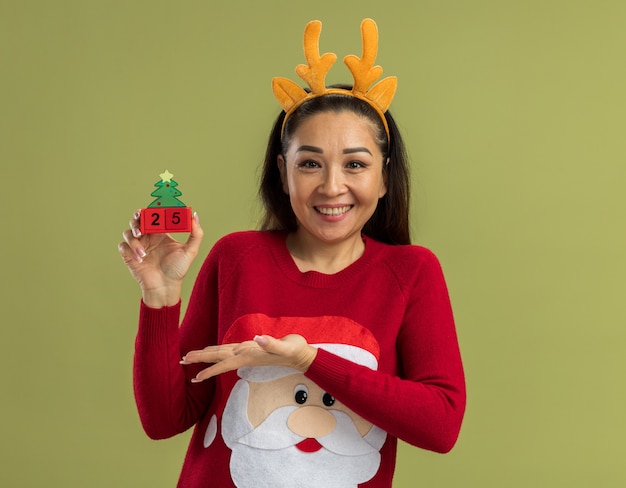 鹿の角と面白い縁を身に着けている赤いクリスマスセーターの幸せな若い女性は、緑の壁の上に元気に立って笑顔で日付25の腕のおもちゃの立方体を提示します