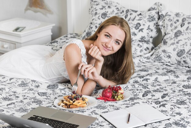 Счастливая молодая женщина, отдыхая на кровати, имея здоровый завтрак