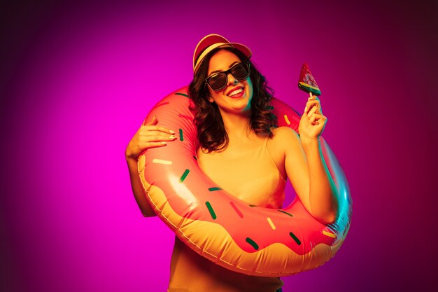 トレンディなピンクのネオンにキャンディーとゴム製のビーチリング、赤いキャップとサングラスで幸せな若い女性