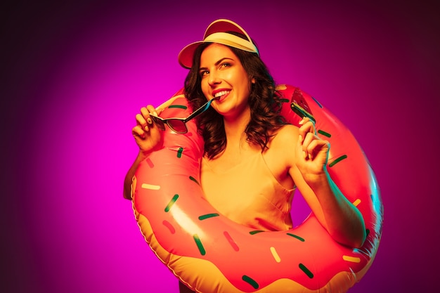 Happy young woman in a rubber beach ring, red cap and sunglasses with a candy on trendy pink neon