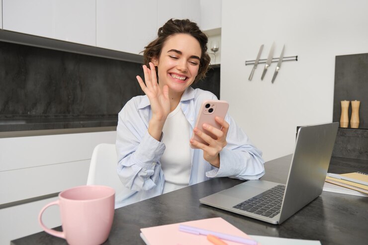 Free Photo happy young woman says hello waves hand at smartphone and smiles video chats with someone on mobile