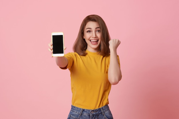 Happy young woman showing at blank screen mobile phone and hand gesture success isolated over pink background.