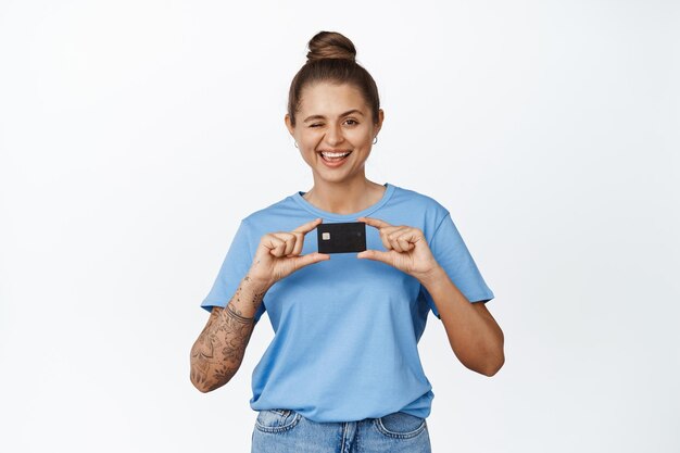 신용 카드를 보여주는 행복한 젊은 여성, 윙크하고 웃고, 파란색 티셔츠를 입은 흰색 배경 위에 서 있는