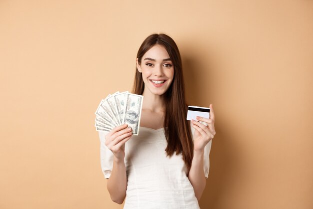 Счастливая молодая женщина, показывающая долларовые купюры и пластиковую кредитную карту, улыбаясь, довольна зарабатыванием денег и покупками ...