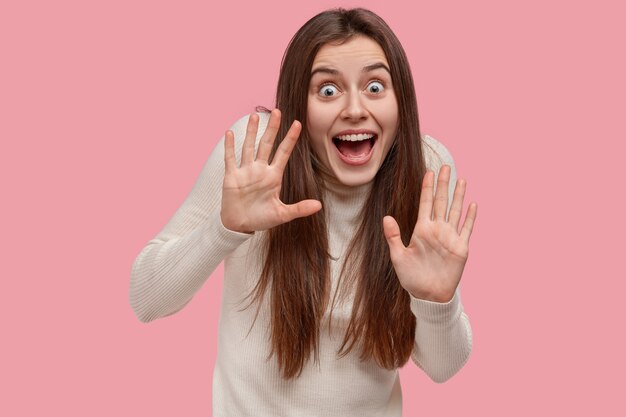 Happy young woman shows palms in protective gesture, feels amazed, impressed by thrilling news, keeps eyes widely opened