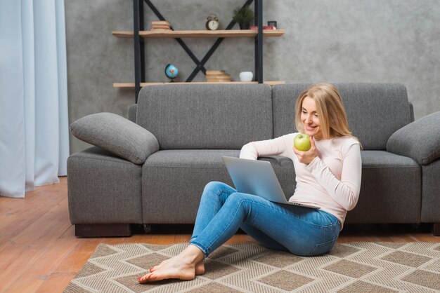 Happy young woman sitting on carpet holding green apple in hand using laptop