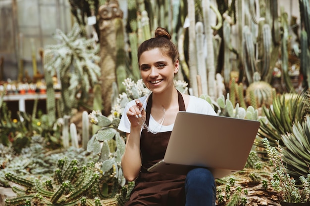 Happy young woman sitting in greenhouse using laptop