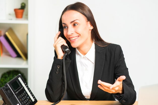 Счастливая молодая женщина, сидя в офисе, разговаривает по телефону жесты