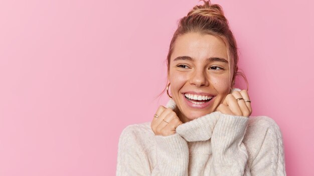 Happy young woman smiles broadly looks gladfully away keeps hands on collar of jumper feels joyful has blonde hair combed in bun stands against pink background copy space for your promotion.
