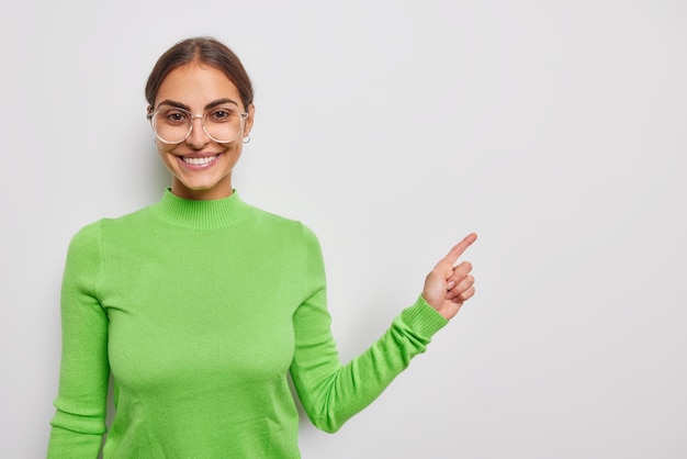 Happy young woman smiles pleasantly wears transparent glasses green turtleneck shows advertisement or promotonal text reads sale banner isolated over white wall