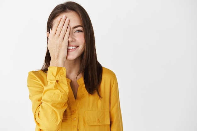 Happy young woman smiling, hiding half of face behind palm, showing one side and staring at camera, checking vision at optician store, standing near copy space on white wall.