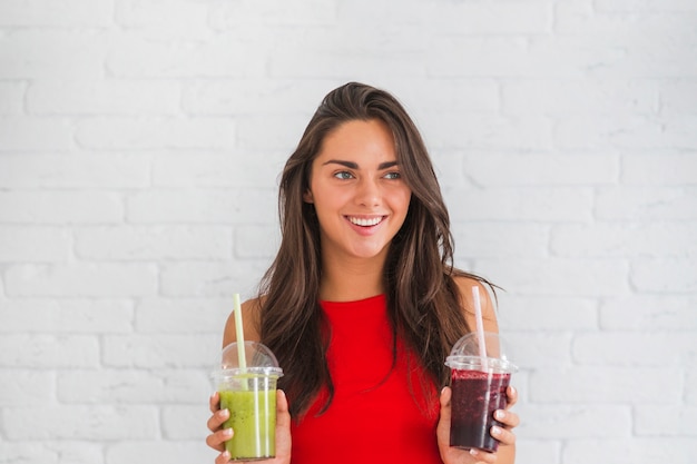 Happy young woman standing against wall holding smoothies in plastic cup