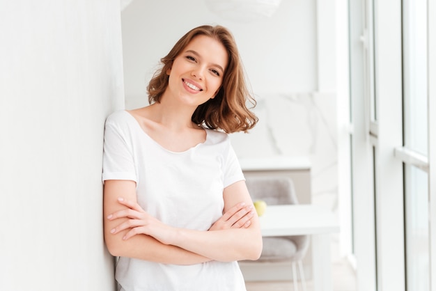 Happy young woman standing near window with arms crossed