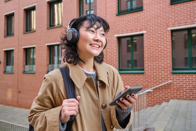 幸せな若い女性がバックパックを持って通りに立ち、スマートフォンがヘッドフォンで音楽を聴いて待っている