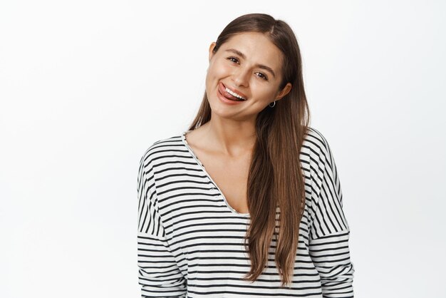 Счастливая молодая женщина приклеивает язык и улыбается, показывая белые здоровые зубы, стоя в полосатой блузке на белом фоне