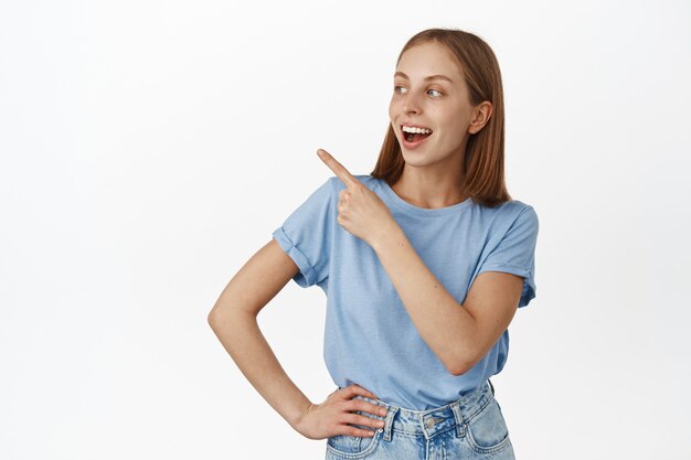 Happy young woman, student pointing and looking at upper left corner with carefree face, laughing over funny advertisement, sale banner, standing over white wall