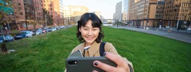 Free photo happy young woman student takes selfie on street holds her tablet and homework notes uses smartphone