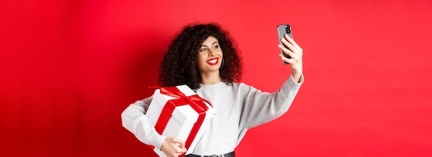 Happy young woman taking selfie with her valentines day gift holding present and photographing on sm