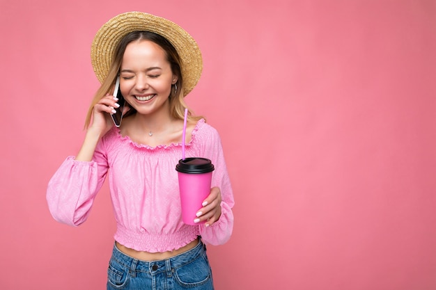Happy young woman talking on mobile phone while having coffee on pink background